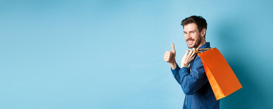 Cheerful shopper holding orange shopping bag on shoulder, turn around at camera with thumbs up, recommending store, blue background.