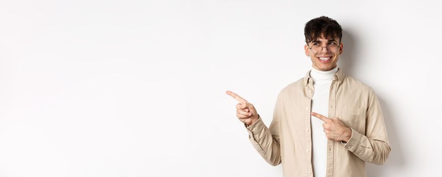 Handsome male model in glasses and trendy clothes, looking and pointing left at empty space, smiling with happy and pleased face expression, white background.