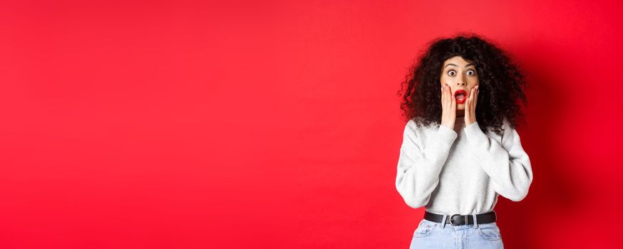 Portrait of shocked woman scream amazed, touching face and looking at camera at impressive promo offer, standing in sweatshirt on red background.