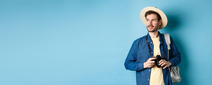 Handsome positive guy in summer hat, holding photo camera and looking aside, tourist taking pictures on vacation, standing with backpack on blue background.