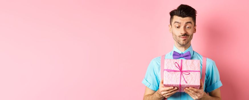 Holidays and celebration concept. Happy smiling guy receive white day gift from his valentine, looking at present with intrigued face, standing over pink background.