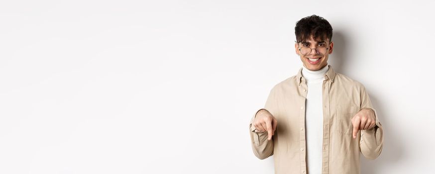 Stylish hipster male model in glasses pointing fingers down, smiling pleased at camera, recommending product, standing on white background.