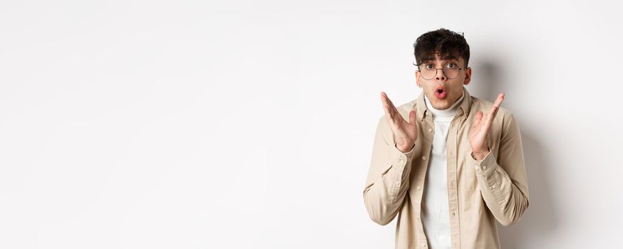 Portrait of surprised hipster guy in glasses checking out awesome promo, say wow and gasping amazed, raising hands up with disbelief, standing on white background.