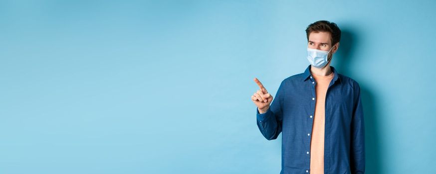 Handsome young man wearing medical mask from coronavirus, pointing finger left and looking satisfied, smiling at empty space, blue background.