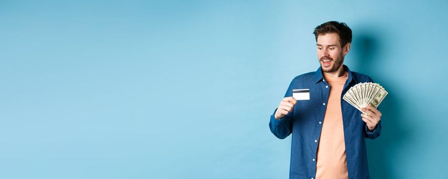 Handsome young man showing cash and looking at plastic credit card, prefer contactless payment, standing on blue background.