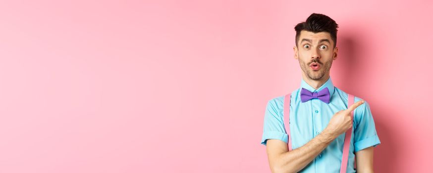 Intrigued and surprised cute guy with moustache showing awesome promo, looking curious and saying wow, pointing finger right at logo, standing on pink background.