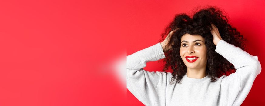 Beauty. Close-up portrait of carefree woman touching her curly hair and looking happy aside at logo, smiling with white teeth and red lips, studio background.