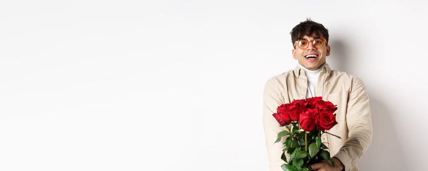 Man in love holding red roses and looking tenderly at camera, staring at lover with happy face, celebrating Valentines day with girlfriend, standing over white background.