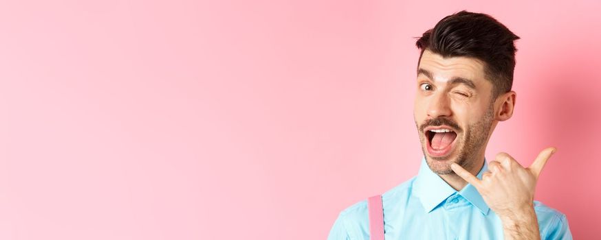 Cheeky and funny guy showing phone gesture, asking to call him and winking, telling dial his number, standing over pink background. Copy space