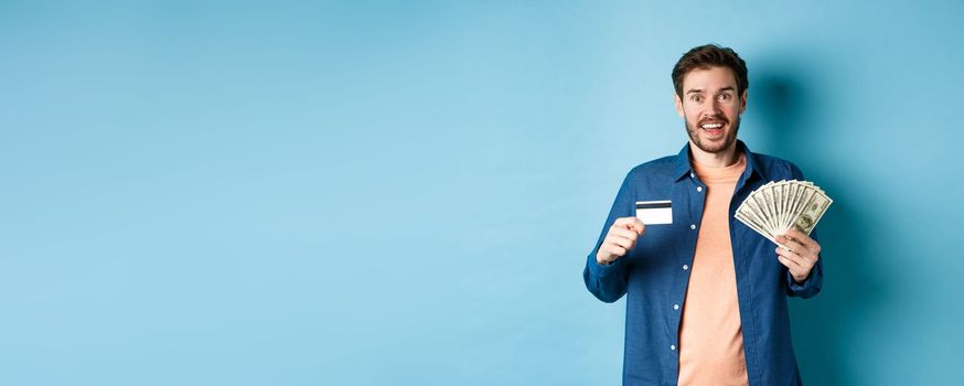 Smiling caucasian guy showing plastic credit card and money, looking happy, standing on blue background.