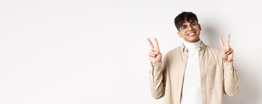 Handsome hipster guy in glasses, smiling with white teeth, showing peace or victory gesture, standing upbeat on white background.