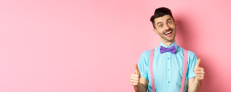 Happy smiling guy in bow-tie and suspenders showing thumbs up, praising good job, approve nice work, standing over pink background satisfied.