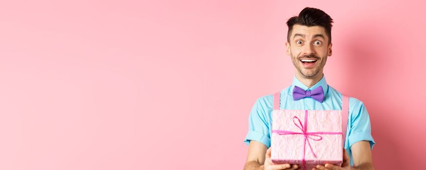 Holidays and celebration concept. Cheerful guy wishing happy birthday and giving you gift wrapped in box, standing over pink background in festive clothes.