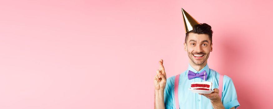 Holidays and celebration concept. Happy young man enjoying birthday party, wearing cone hat and cross fingers, making wish on bday cake with candle, pink background.