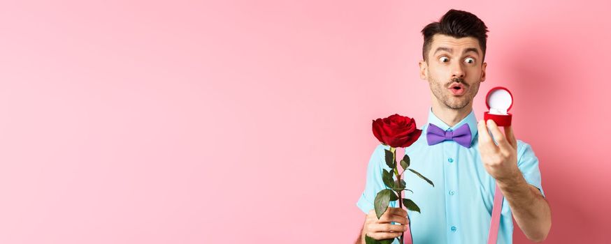 Valentines day. Romantic funny guy going to make wedding proposal, asking to marry him, holding red rose and looking at engagement ring with excitement, pink background.