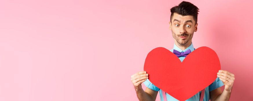 Valentines day concept. Cute boyfriend looking at big red heart, waiting for lover on romantic date, pink background.