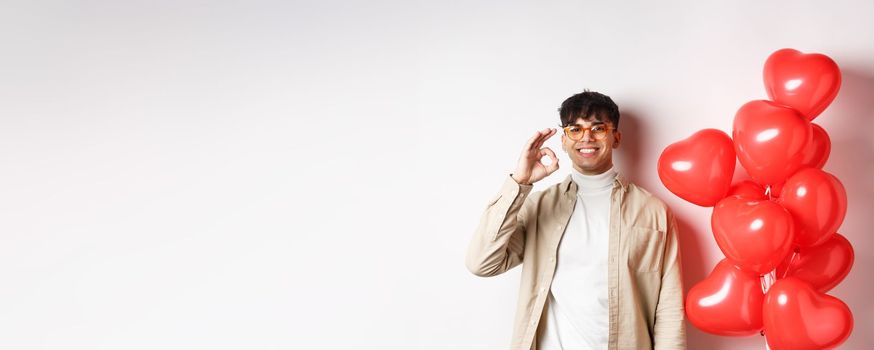 Valentines and romance concept. Happy young man smiling, showing OK sign near red hearts gesture, recommending good offer on lovers day.