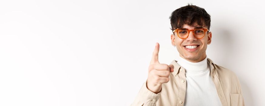 Close-up of lucky handsome man in glasses, smiling and pointing finger at camera, praising something good, standing on white background.