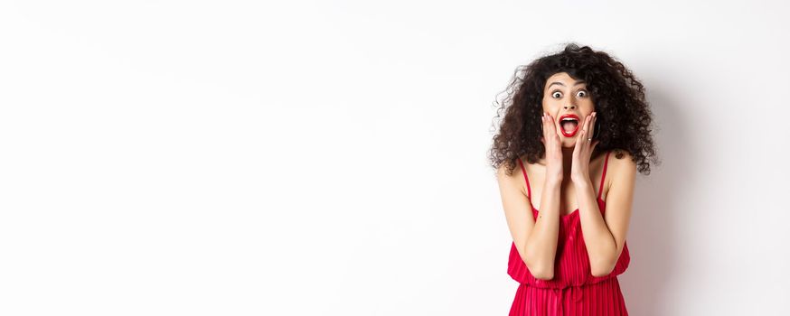 Image of elegant woman scream surprised, staring at camera, see promo offer and shouting of joy, standing in dress over white background.