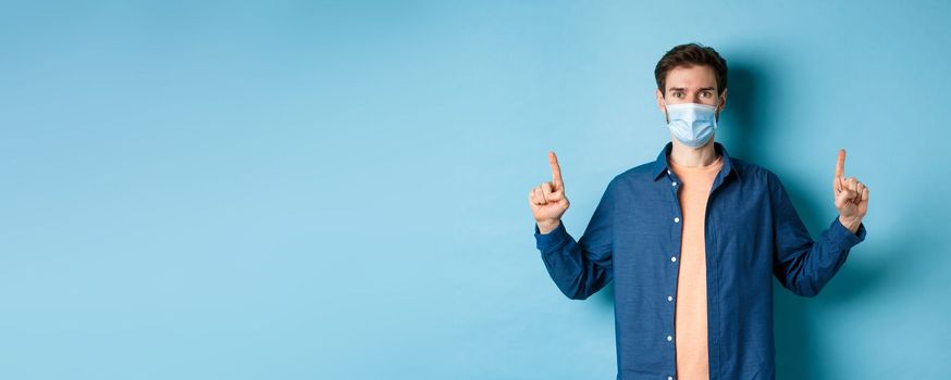 Covid-19 and lifestyle concept. Handsome young man in face mask looking healthy and happy, pointing fingers up at empty space, blue background.