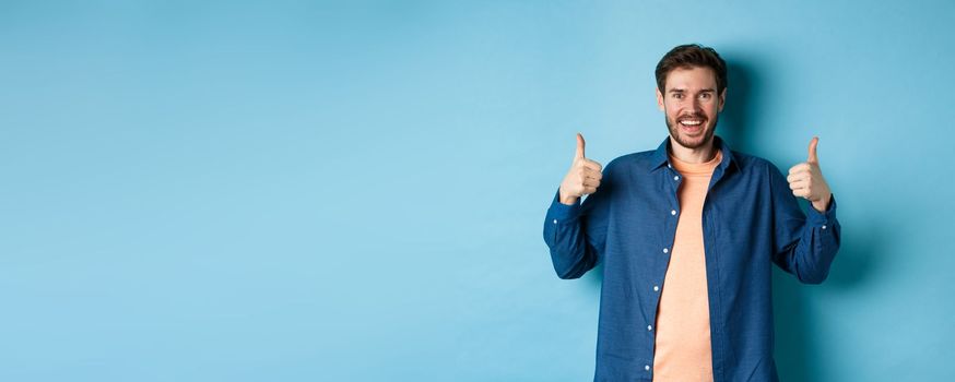 Cheerful caucasian guy in casual shirt showing thumbs up in approval, smiling and praising something good, standing on blue background.