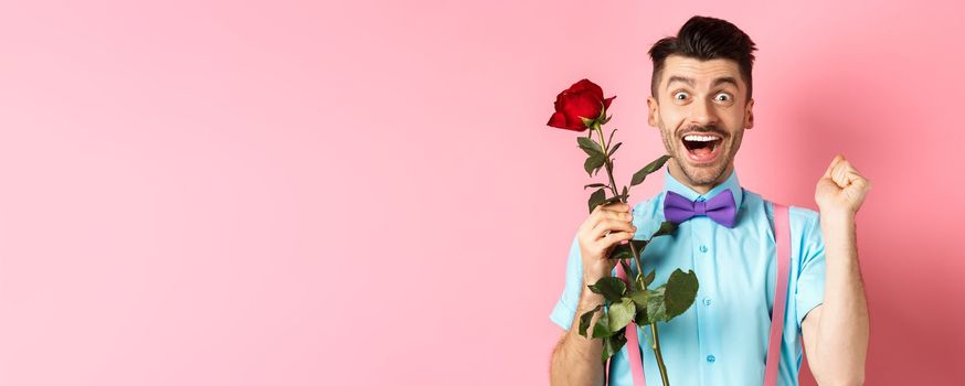 Valentines day and romance concept. Excited boyfriend jumping from happiness on romantic date, holding red rose and celebrating, standing on pink background.