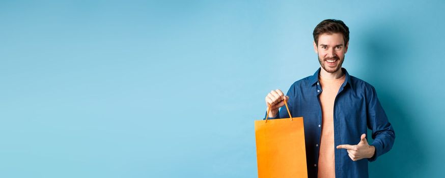 Smiling caucasian man pointing finger at orange shopping bag with bought items, standing on blue background.