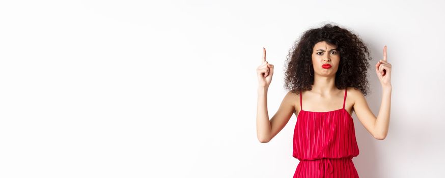 Disappointed frowning woman with makeup, wearing red dress on valentines, pointing fingers up and complaining, standing on white background.