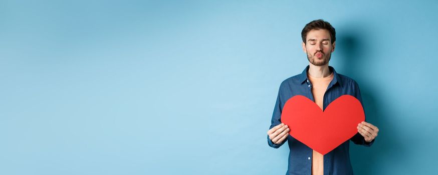 Romantic boyfriend with valentines red heart close eyes, pucker lips and waiting for kiss on lovers day, standing against blue background.