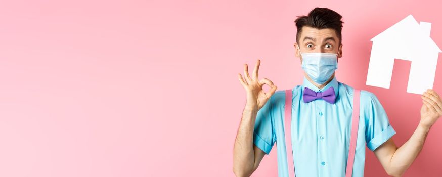 Covid, pandemic and real estate concept. Excited young man in medical mask showing OK sign and paper house cutout, recommending agency, standing over pink background.