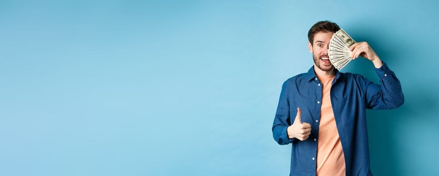 Cheerful guy cover half of face with money and showing thumbs-up, recommending fast cash loan, standing on blue background.