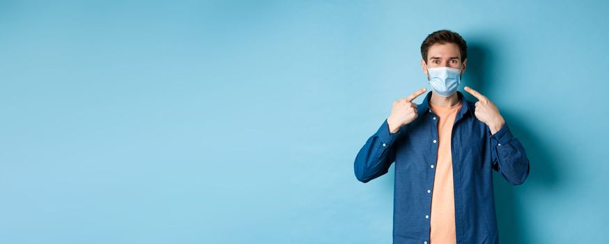 Coronavirus and pandemic concept. Portrait of happy and healthy man pointing fingers at his medical mask, smiling at camera, standing on blue background.