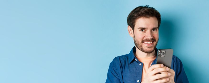 Close up of young guy with beard, smiling happy at camera and holding mobile phone, standing on blue background.