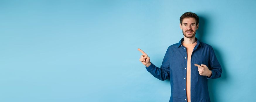 Happy smiling caucasian man showing advertisement, pointing fingers left at empty space, standing on blue background.