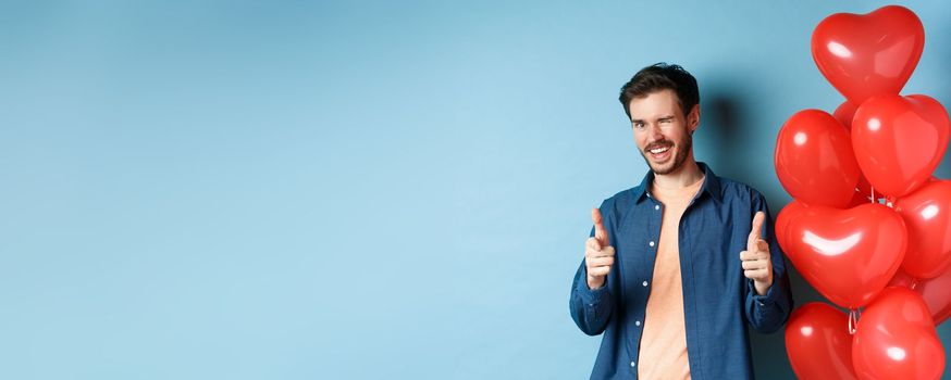 Happy valentines day. Cheeky boyfriend winking and pointing fingers at camera, standing with red romantic balloons on blue background. Copy space