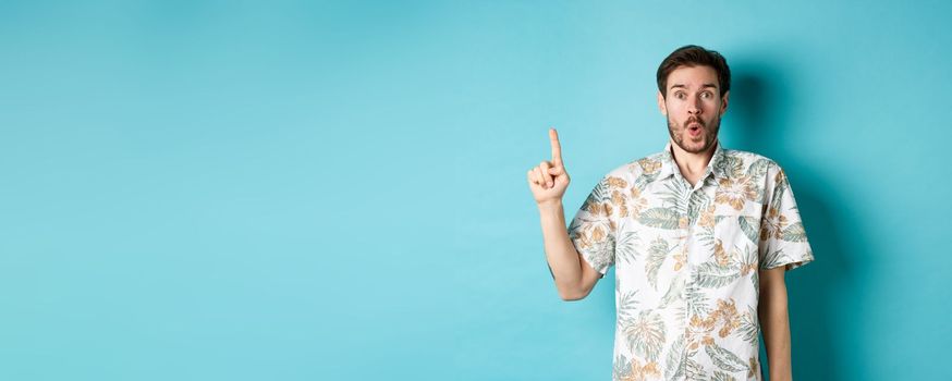 Surprised caucasian tourist in hawaiian shirt, pointing finger up and say wow, checking out promotion, standing on blue background.