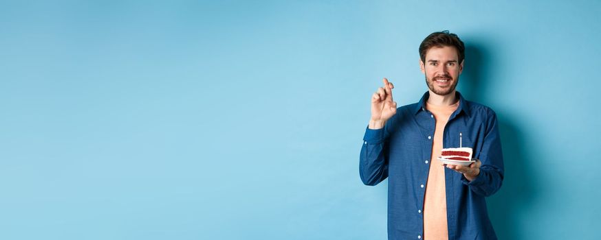 Celebration and holiday concept. Hopeful young man making wish on birthday cake, cross fingers for good luck and smiling positive, standing on blue background.