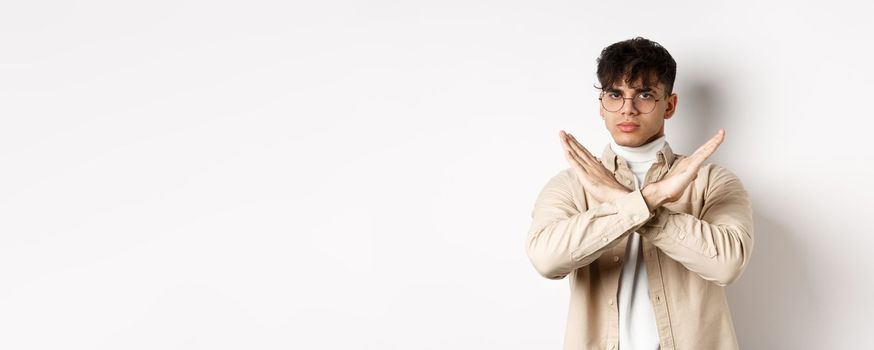 Image of serious young guy in glasses say no, showing cross gesture, make stop sign to disagree or prohibit something, standing on white background.