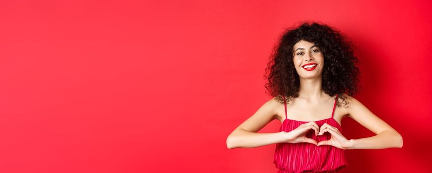 Lovers day. Beautiful woman celebrating valentines, showing heart sign and smiling, standing in romantic red dress on studio background.