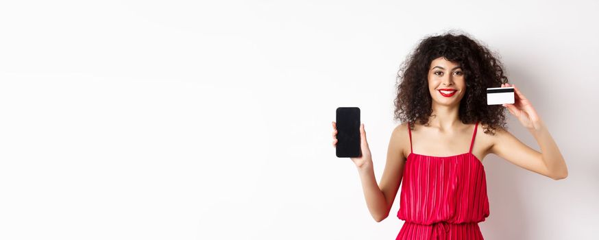 Online shopping concept. Elegant female model with curly hair, wearing red dress, showing plastic credit card with empty mobile phone screen, standing on white background.