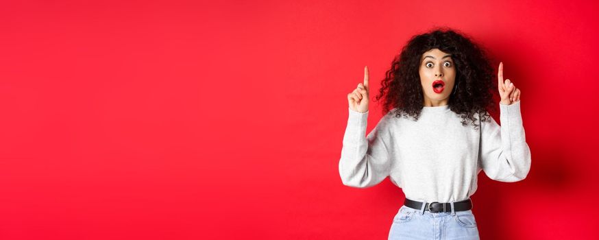 Startled girl showing amazing news, drop jaw and gasping surprised as pointing fingers up at empty space, standing in sweatshirt and jeans on red background.