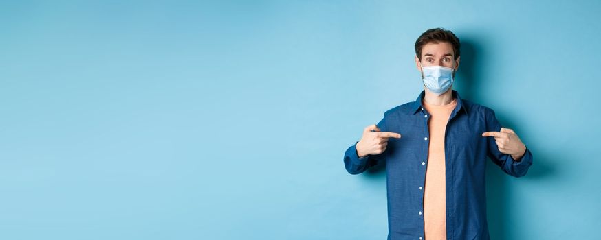 Covid-19 and pandemic concept. Healthy young man in medical mask pointing fingers at center logo, standing on blue background.