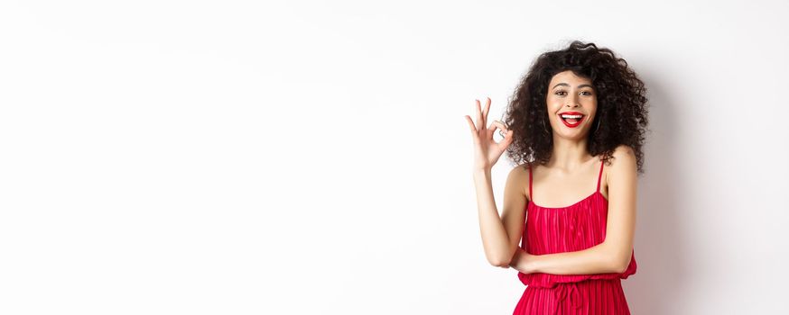 Cheerful caucasian woman with curly hair and makeup, wearing elegant red dress, showing OK sign and smiling in approval, standing over white background.