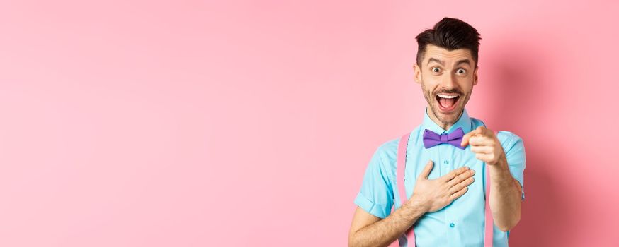 Funny guy laughing at something hilarious, smiling amazed and pointing finger at camera, standing on pink background.