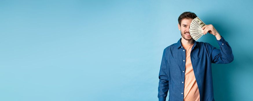 Happy young guy covering half of face with dollars and smiling, winning prize money, standing on blue background.