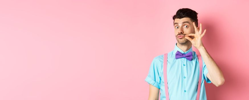 Funny young man touching his french moustache and pucker lips, looking silly at camera, standing in bow-tie and suspenders on pink background.