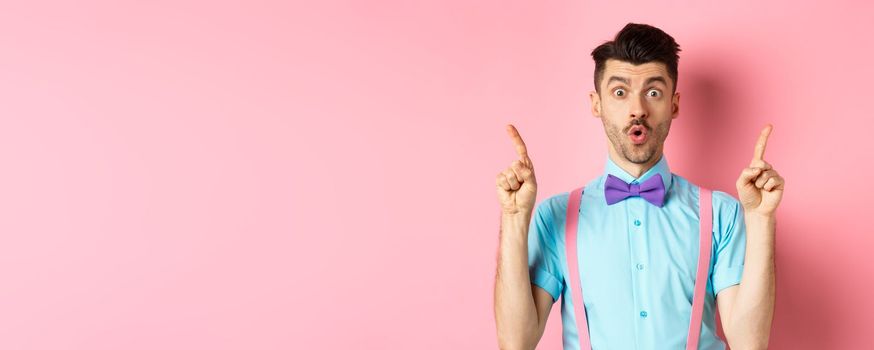 Impressed young guy with bow-tie point fingers up, showing top promo offer and say wow, standing over pink background.