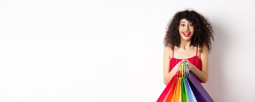 Cheerful young woman in stylish red dress, holding shopping bags and smiling, buying during promo offers, standing over white background.