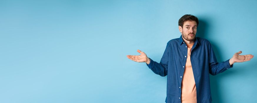 Confused young man with beard, shrugging shoulders and looking clueless at camera, know nothing, standing on blue background.