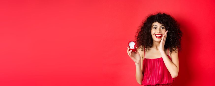 Image of surprised young woman with curly hairstyle, wearing red dress and lipstick, showing engagement ring, going to get married, standing on studio background.